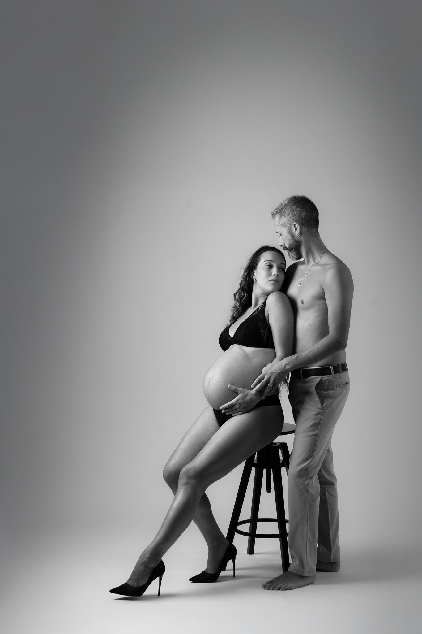 Black and white photo of a pregnant woman in lingerie sitting on a stool with her shirtless partner standing behind her.