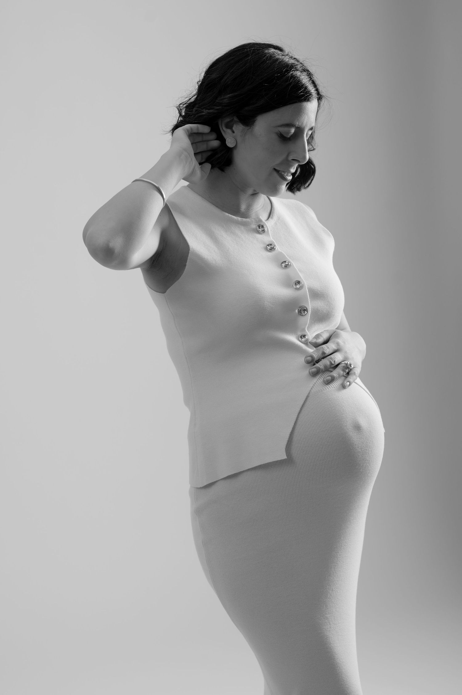 Black and white maternity photo of a pregnant woman in a white outfit, elegantly posing while cradling her baby bump.