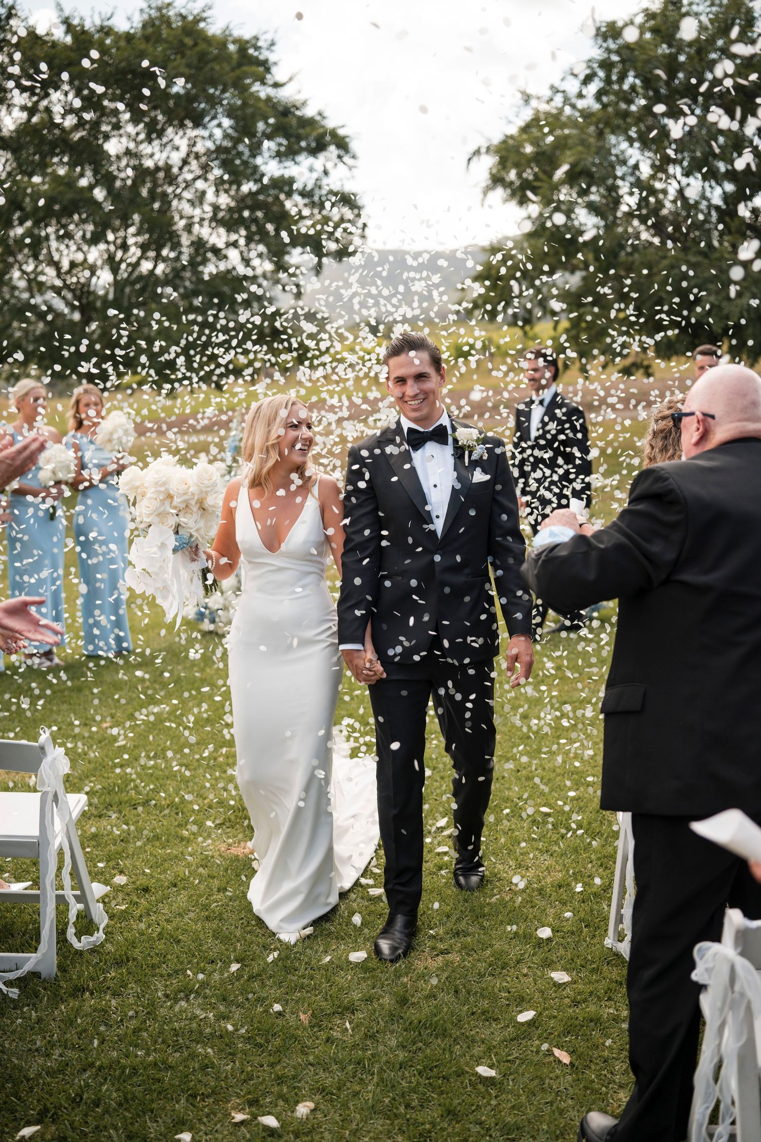 Bride and groom walk down aisle as guests toss white flower petals at outdoor wedding ceremony.