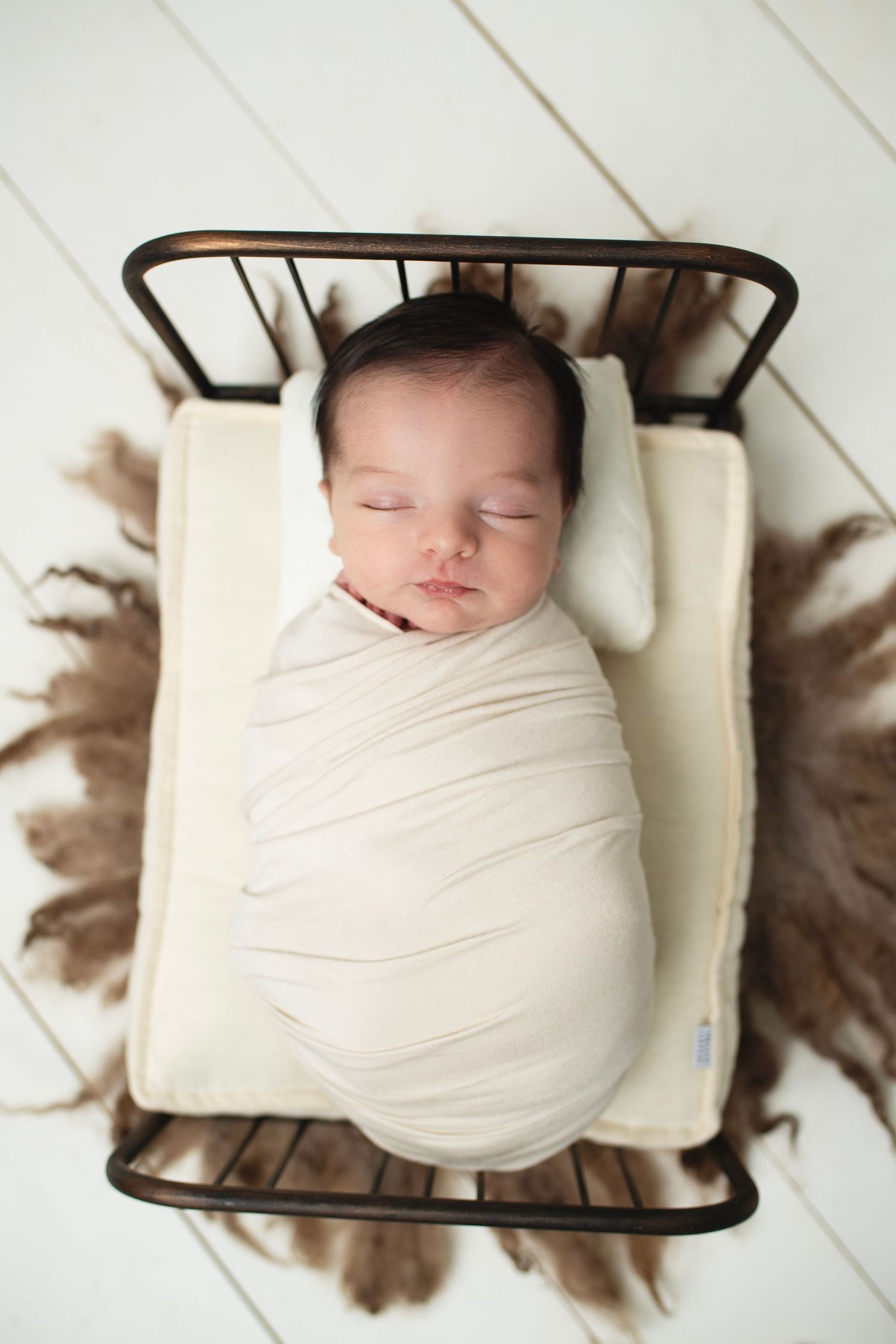 Swaddled newborn baby sleeping in a small metal bed with fur blanket.
