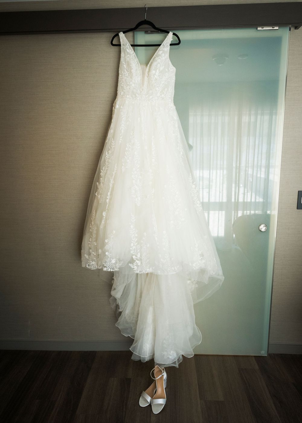 Elegant white lace A-line wedding gown hanging on a door with white bridal heels below.