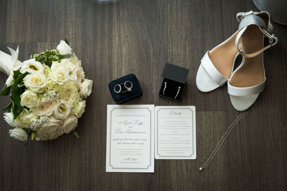 Wedding flat lay with white rose bouquet, gold rings, pearl earrings, white heels, and elegant invitations on wood floor.