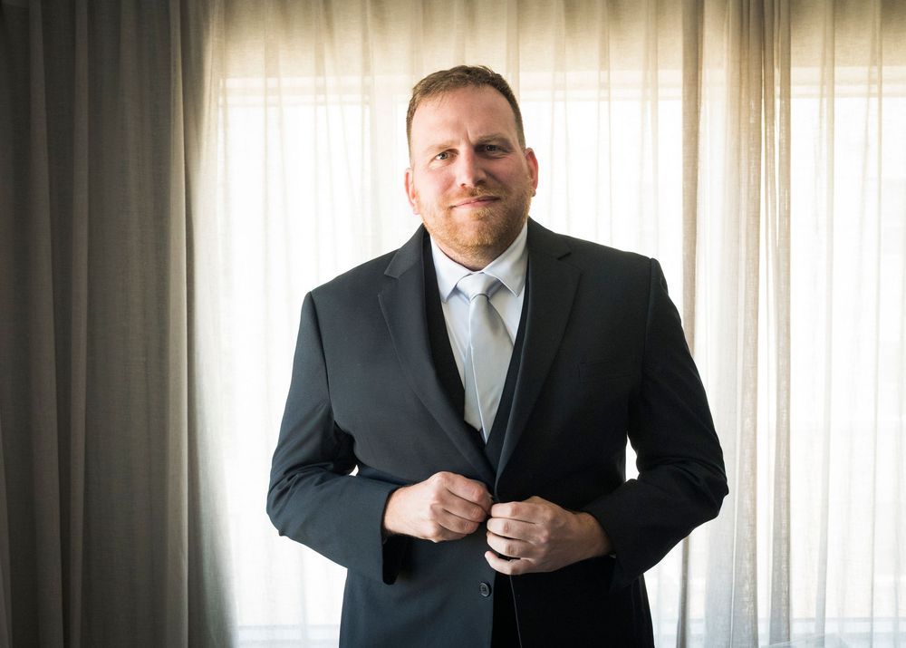 Man in dark suit adjusting jacket buttons, standing near sheer curtains in natural light.