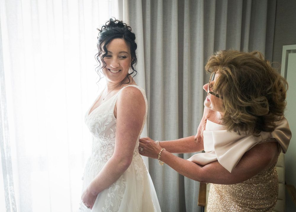 A bride smiles as an older woman helps fasten her white lace wedding gown near a bright window.