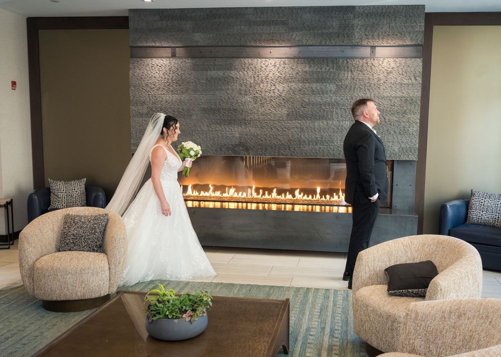 Bride and groom share a first look moment by a modern fireplace in an elegant hotel lobby.