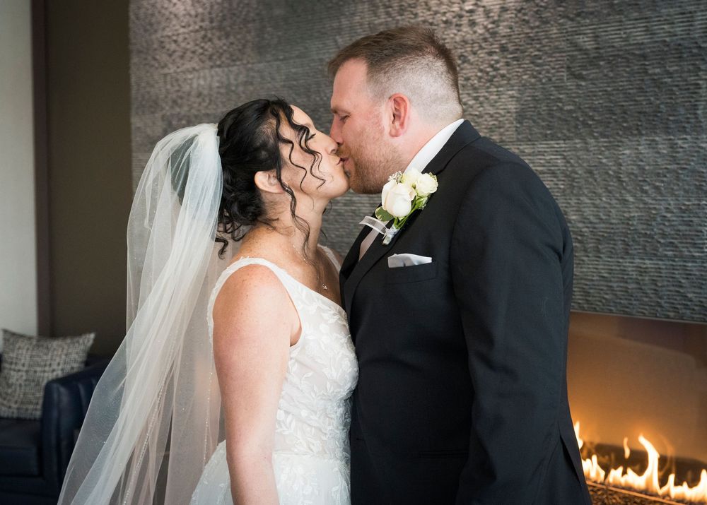 Bride and groom sharing a first kiss in wedding attire near a modern fireplace.