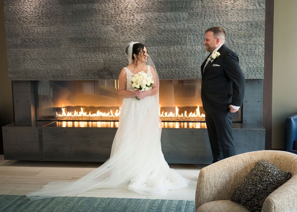 Bride and groom standing together in front of a modern fireplace, bride holding white bouquet in elegant wedding attire.