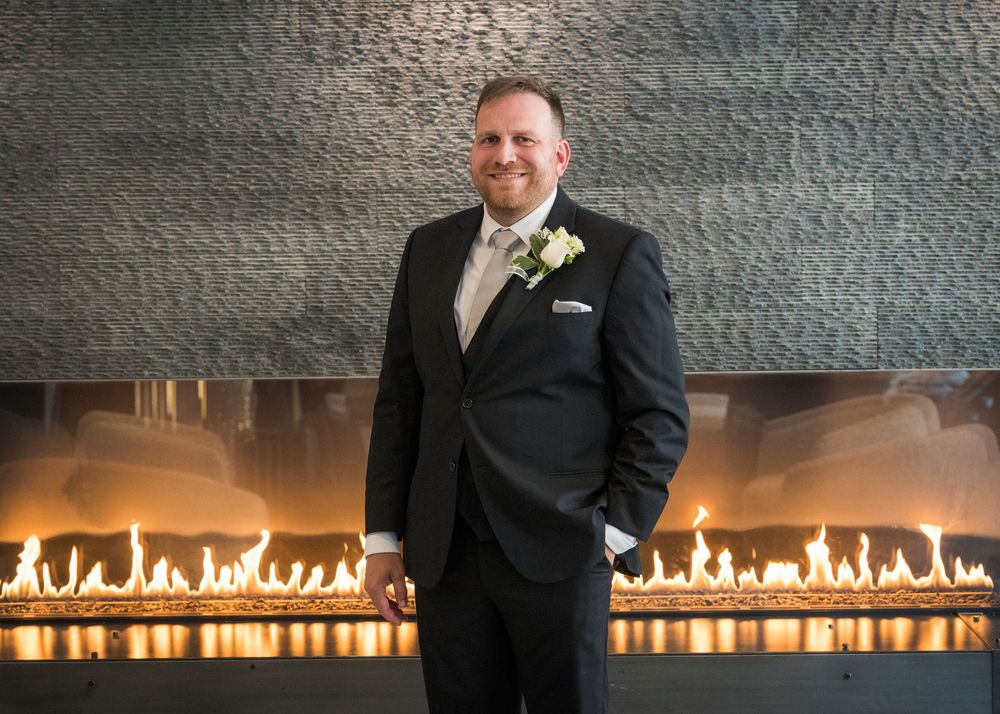 Smiling groom in black tuxedo with white boutonniere standing before a modern linear fireplace.