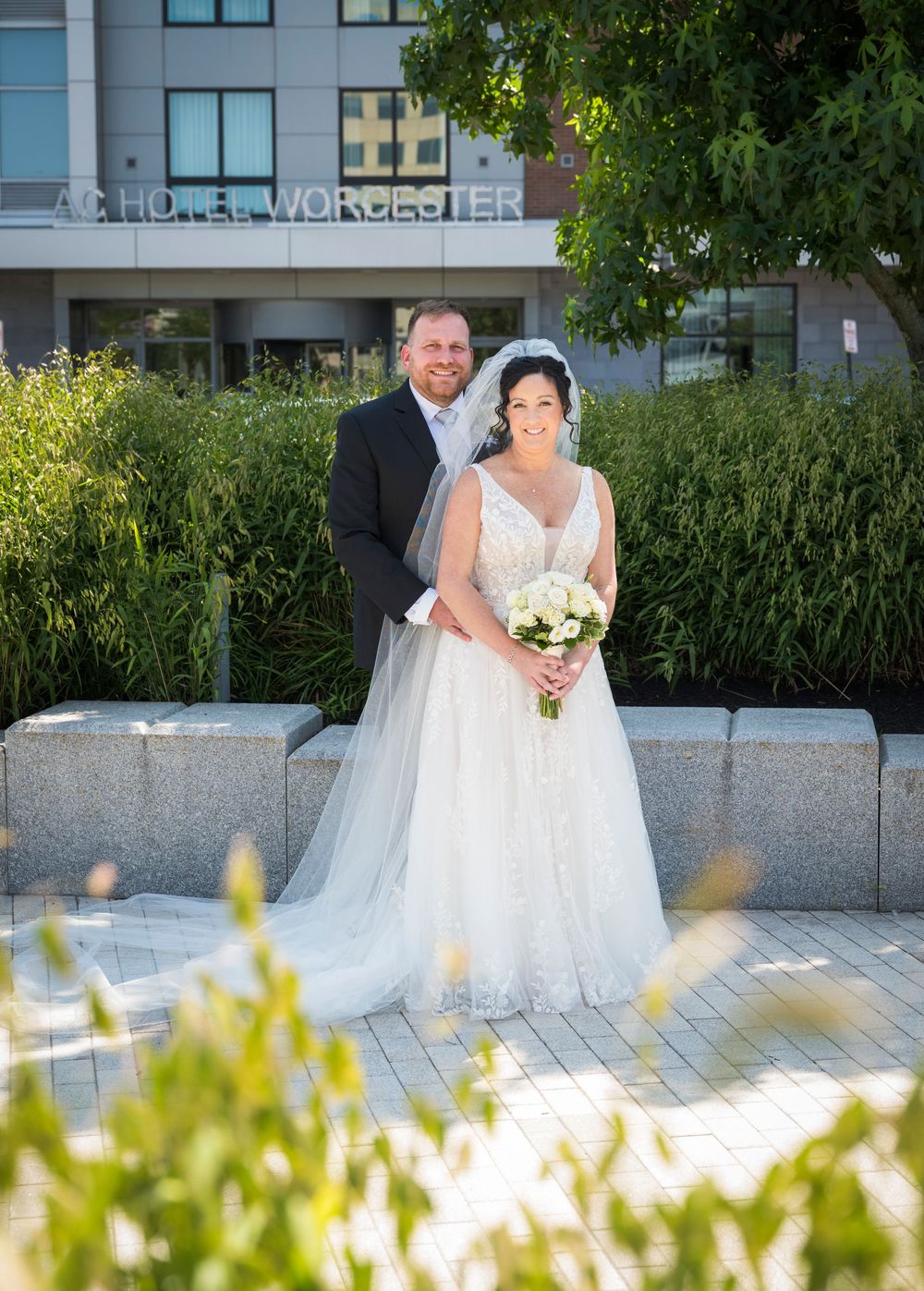 Bride and groom posing outdoors at AC Hotel Worcester on their wedding day, bride holding white bouquet.
