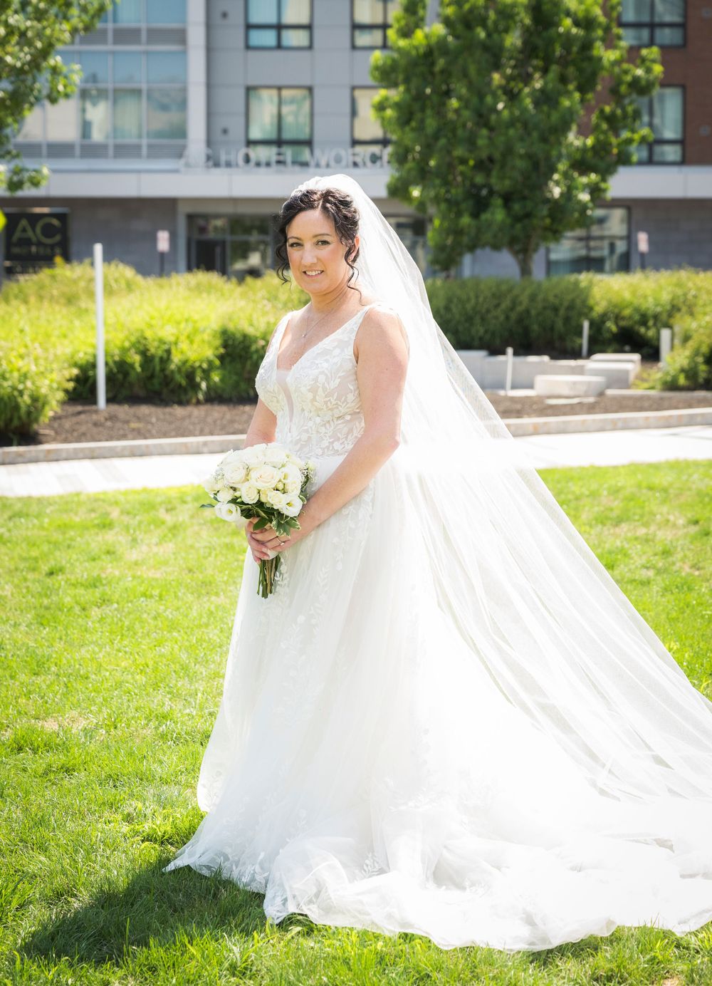 Bride in white lace ball gown and veil holds bouquet on green lawn with modern building backdrop.