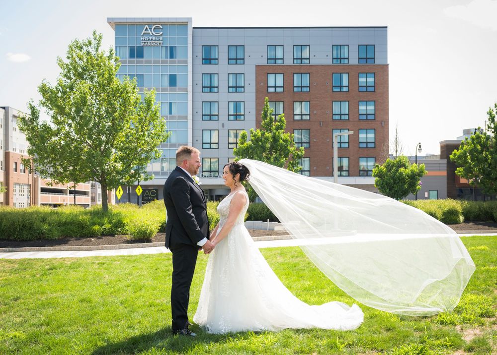 Bride and groom share romantic moment outdoors, veil flowing in wind, AC Hotel in background.