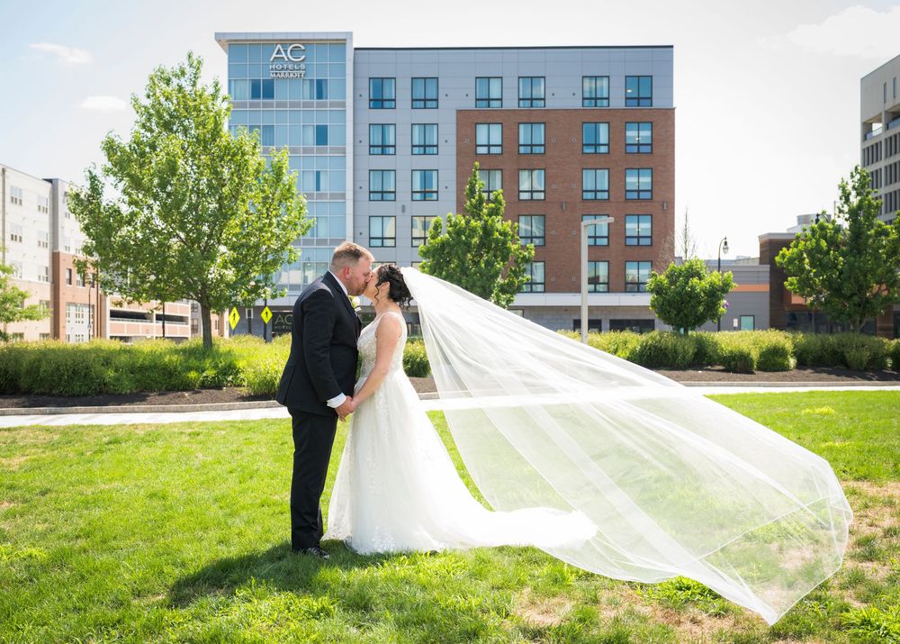 Bride and groom kiss on green lawn, cathedral veil billowing, AC Hotel visible behind them.