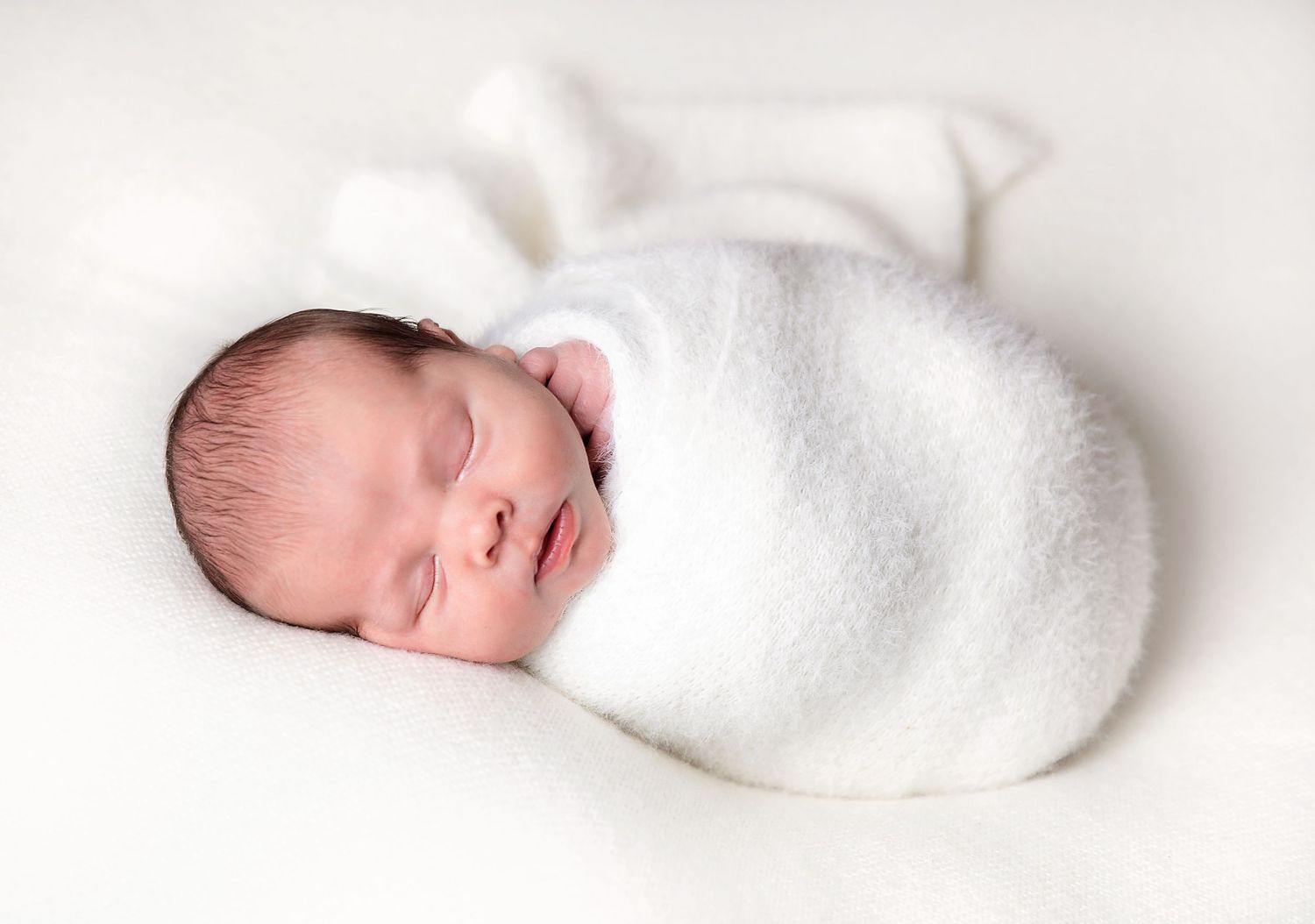 Peaceful newborn baby sleeping wrapped in a soft white blanket, with dark hair and delicate features. During a newborn session in Orpington
