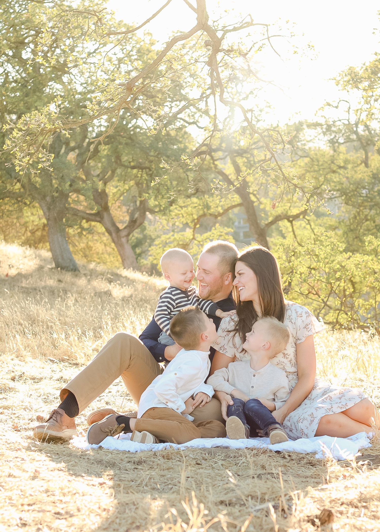 Family of five sitting on a blanket outdoors in golden sunlight, parents smiling with three young children among trees.