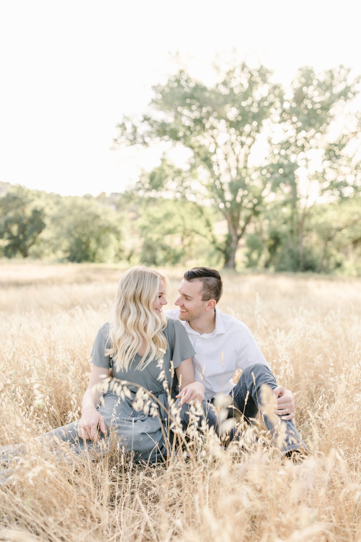 A couple sits together in a golden field, gazing at each other surrounded by tall dry grass and trees.