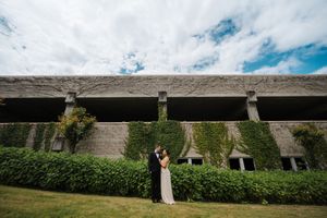 Couple shares romantic moment on green lawn beside ivy-covered stone building under blue cloudy sky.