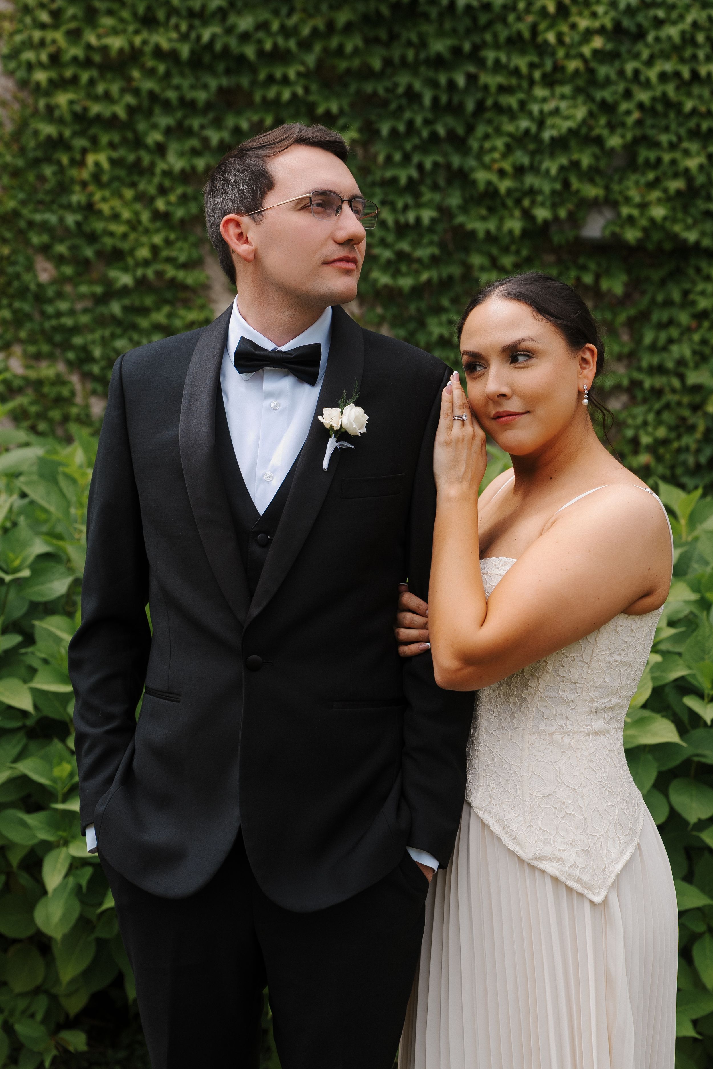 Bride in white gown holds groom's arm in black tuxedo before ivy-covered wall.