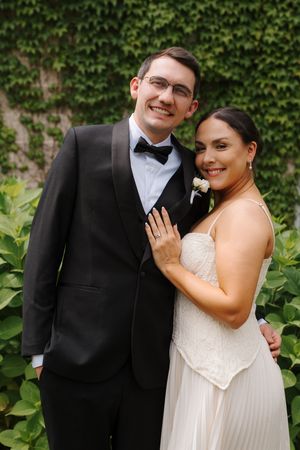 Smiling bride and groom pose together in wedding attire before a lush ivy wall.