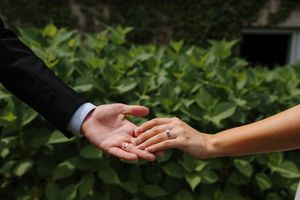 Two hands reaching toward each other, one wearing an engagement ring, with lush green ivy leaves in the background.