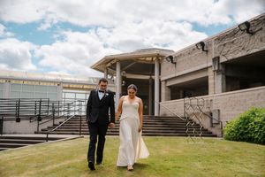 Bride and groom walk hand in hand on green lawn outside modern wedding venue under partly cloudy sky.