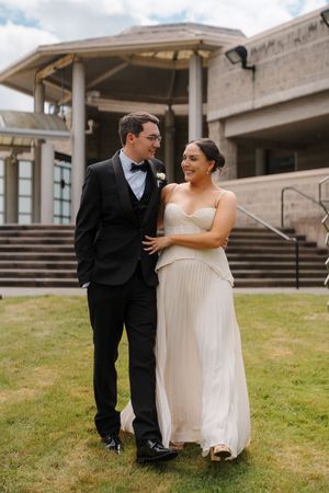 A bride and groom walk together on a lawn outside a stone building, smiling at each other on their wedding day.