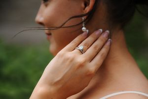 Bride wearing a diamond engagement ring and pearl drop earrings, gently touching her neck against a green background.