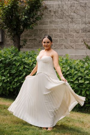 Woman in white pleated maxi dress twirling outdoors with lush green hedges and stone wall backdrop.