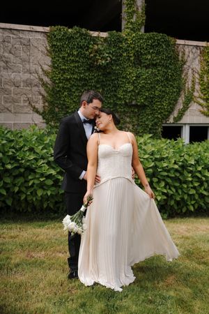 A bride in a flowing white gown holds a bouquet while a groom in a tuxedo embraces her outdoors near an ivy-covered wall.