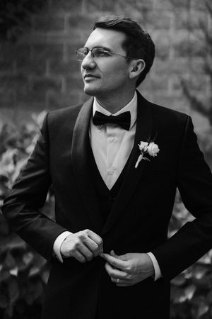 Black and white portrait of a groom in tuxedo with bow tie and boutonniere, adjusting jacket outdoors.