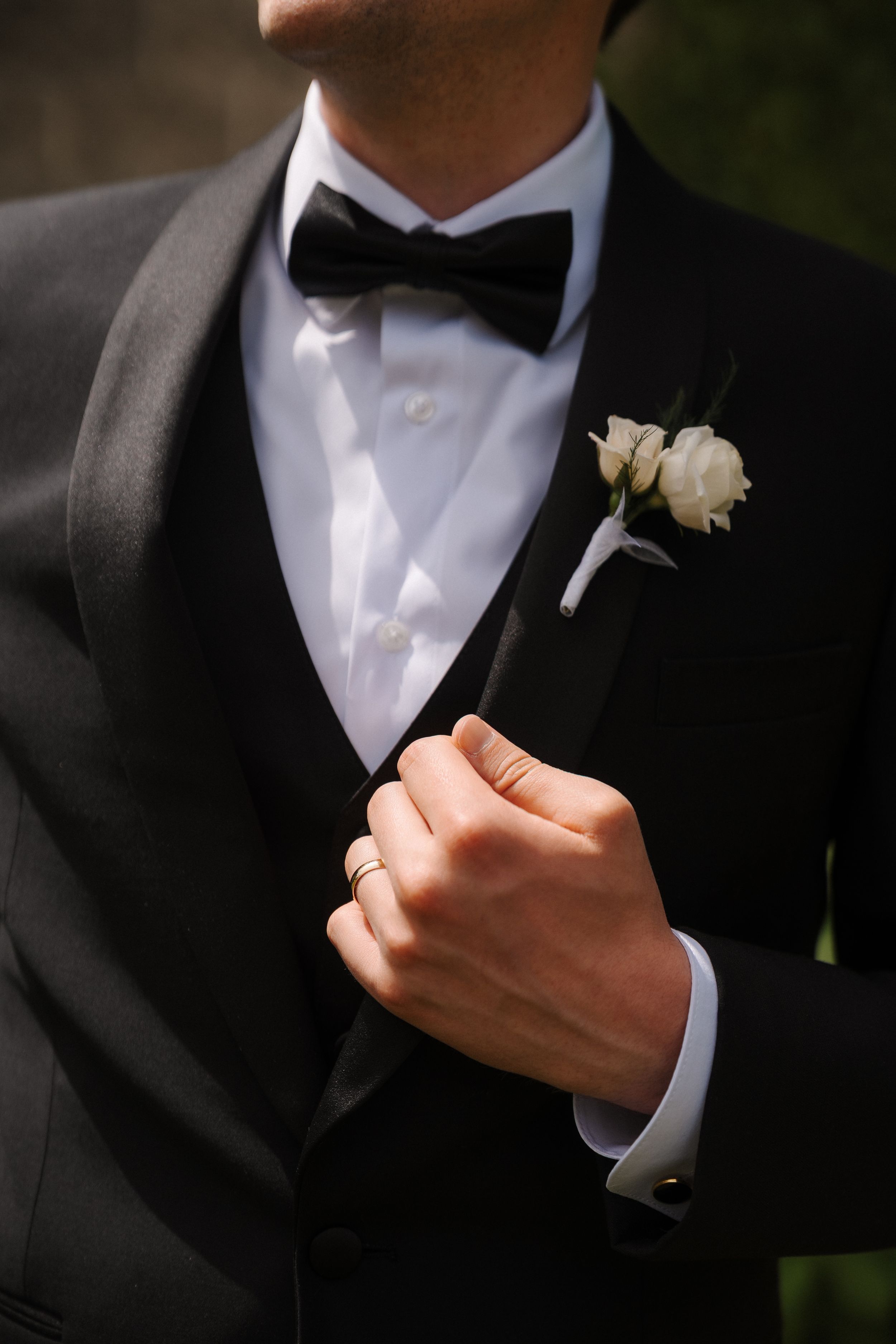Groom in black tuxedo with bow tie and white rose boutonniere, adjusting jacket with ringed hand.