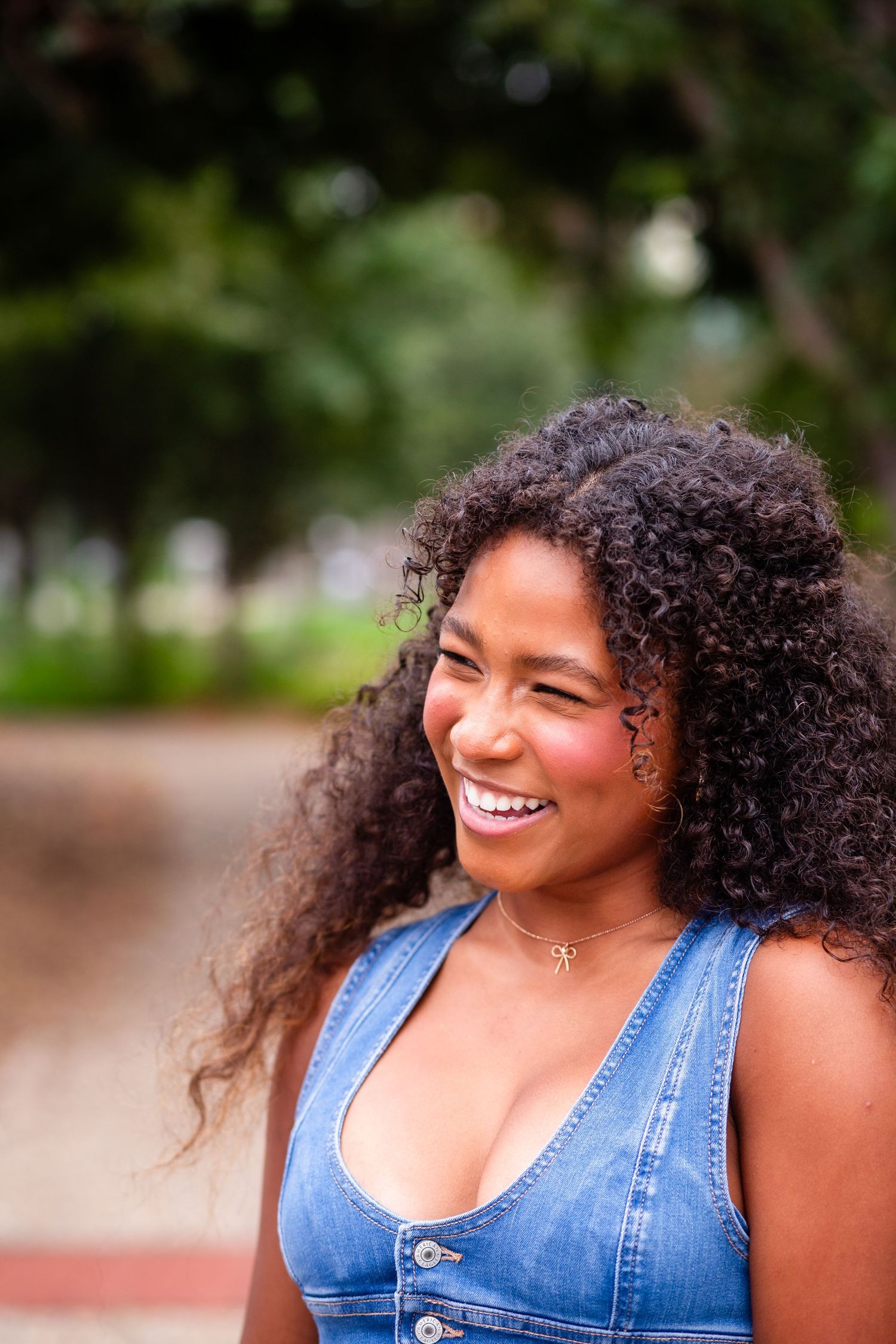 Smiling young woman with curly hair wearing a denim top, laughing outdoors in a park setting.