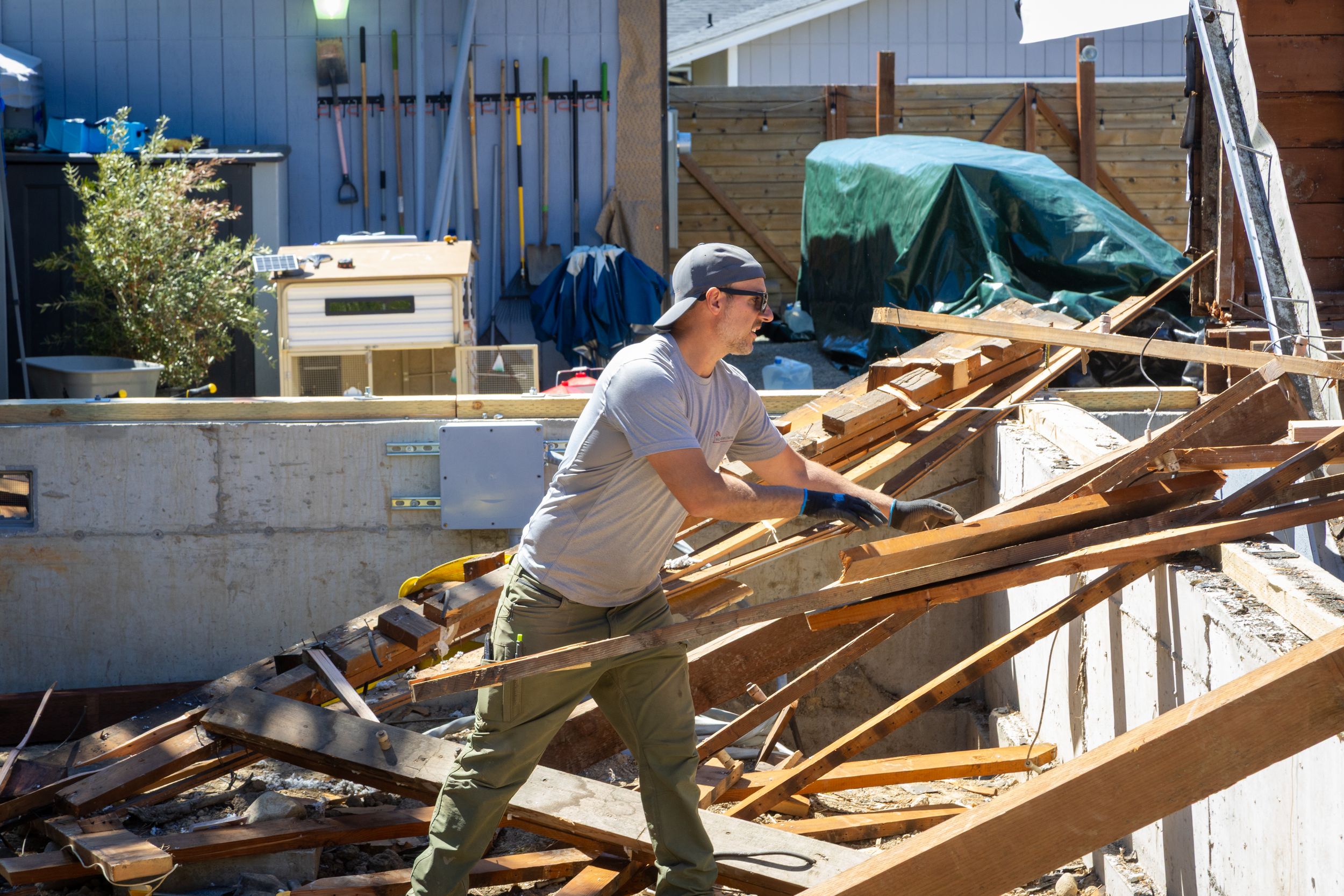 Man in cap and gray shirt clearing wooden debris at a construction or demolition site outdoors.