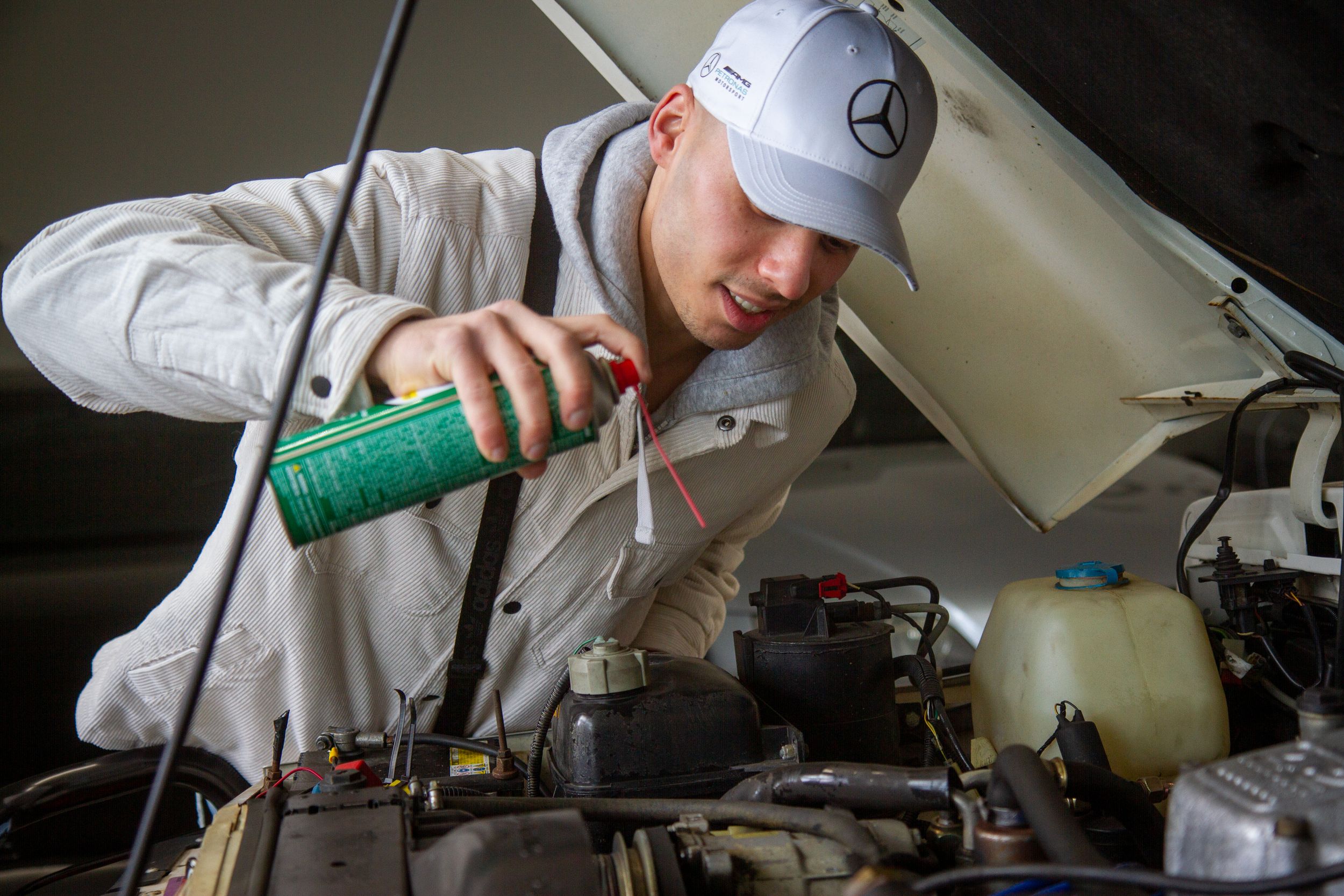 Mechanic in Mercedes-Benz cap pouring green fluid into a car engine under an open hood.