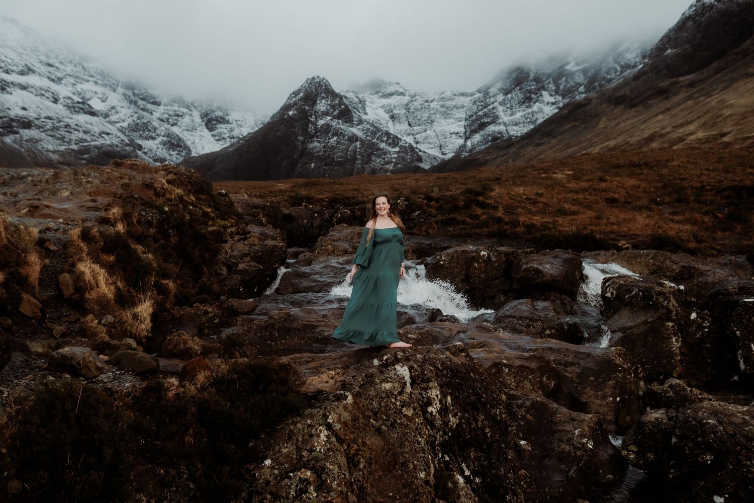 Woman in green gown stands on rocky terrain with snowy mountains and misty skies in Scottish Highlands.