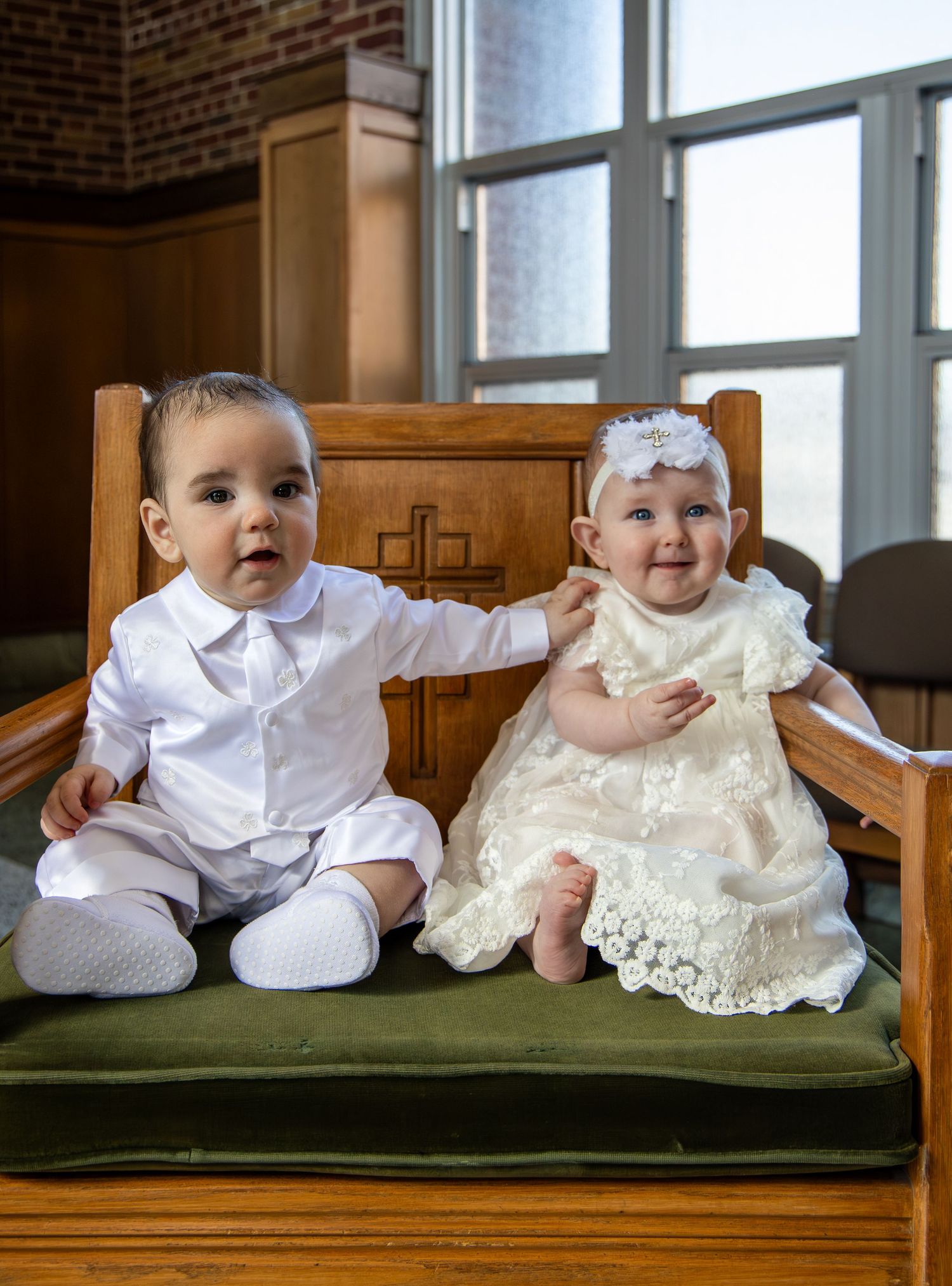 Two babies in white and cream baptismal outfits sitting on a green church pew.