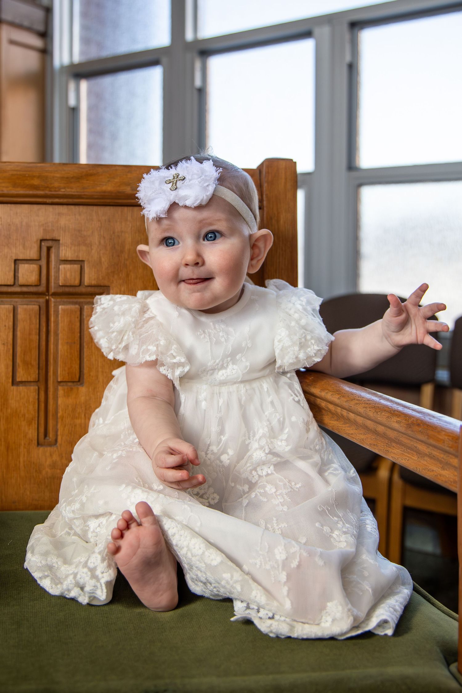 Baby in ornate white lace baptismal gown posing on green church bench.
