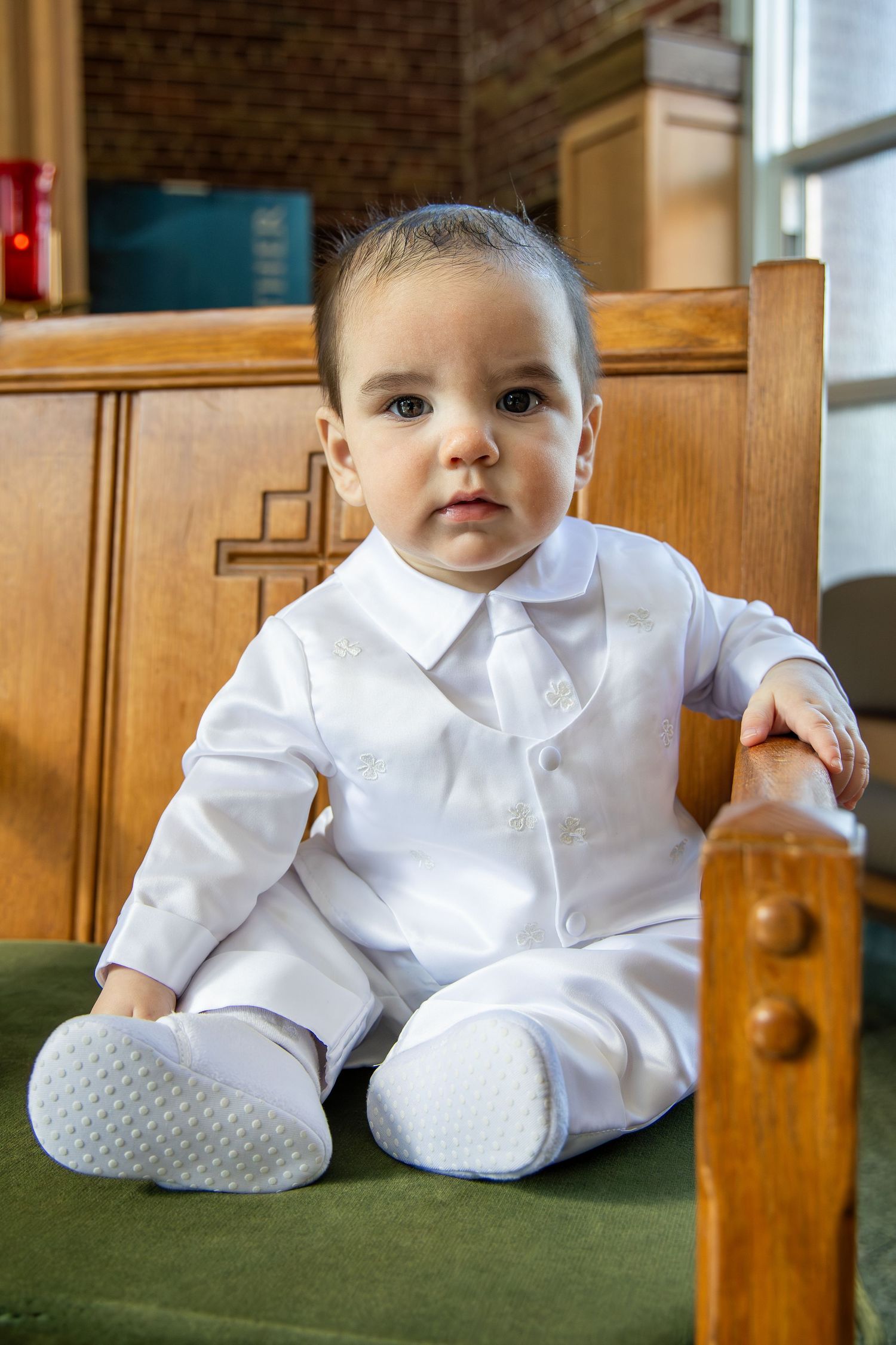 Baby wearing traditional white baptismal outfit sitting on wooden church pew.