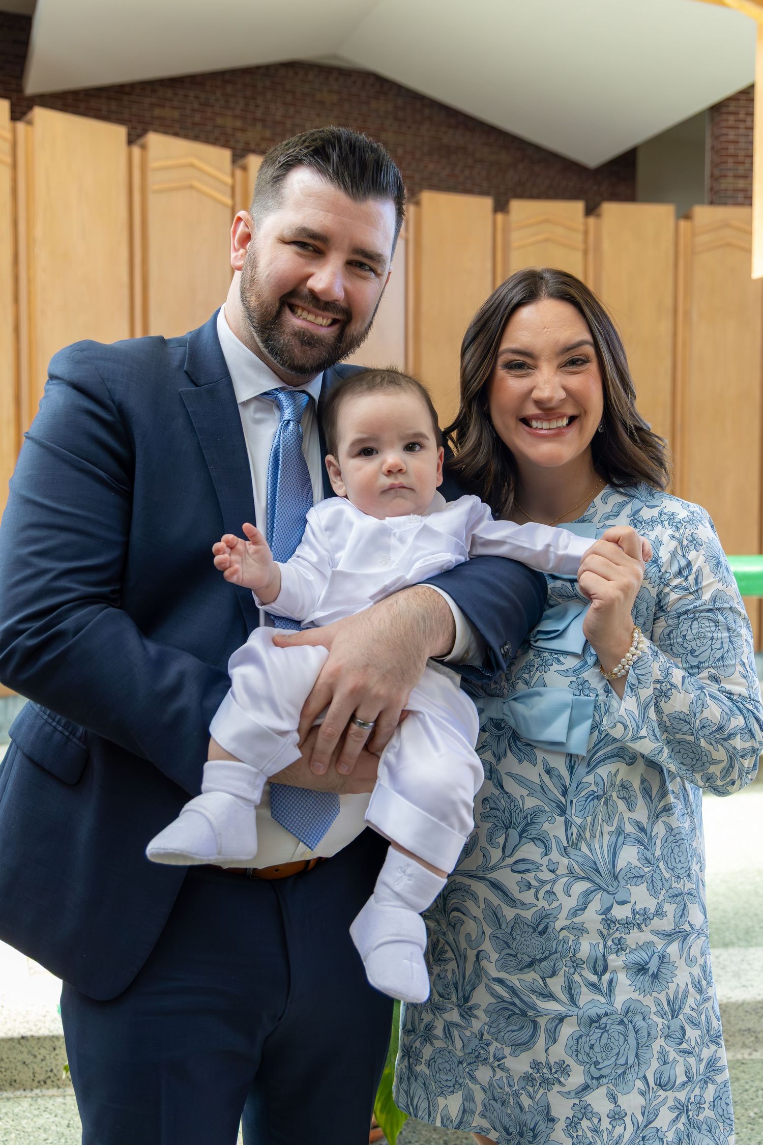 A couple in formal attire shares joyful laughter while holding a baby in white clothing outdoors.