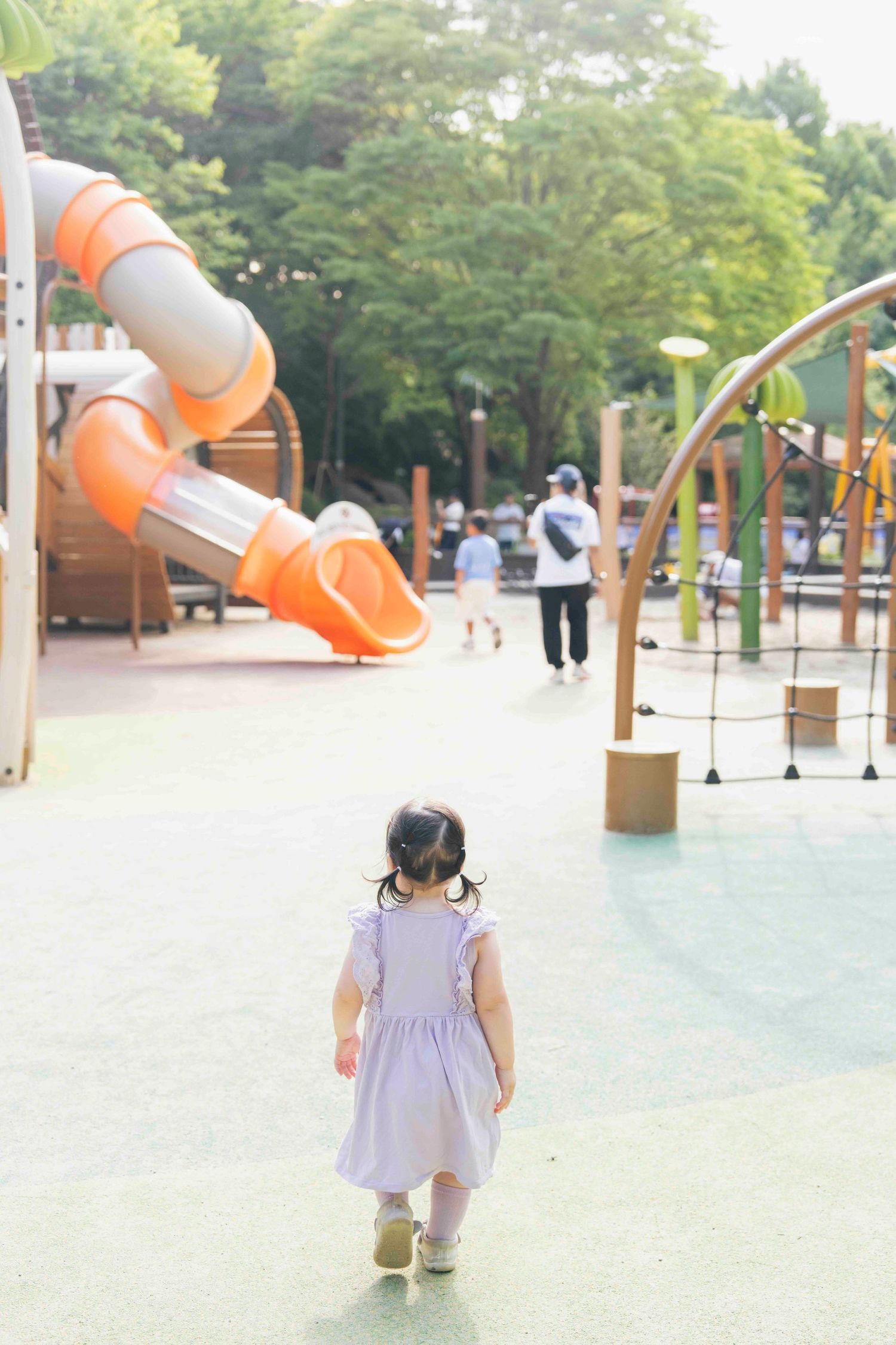 Young toddler girl in purple dress walking toward colorful playground equipment in a sunny park.
