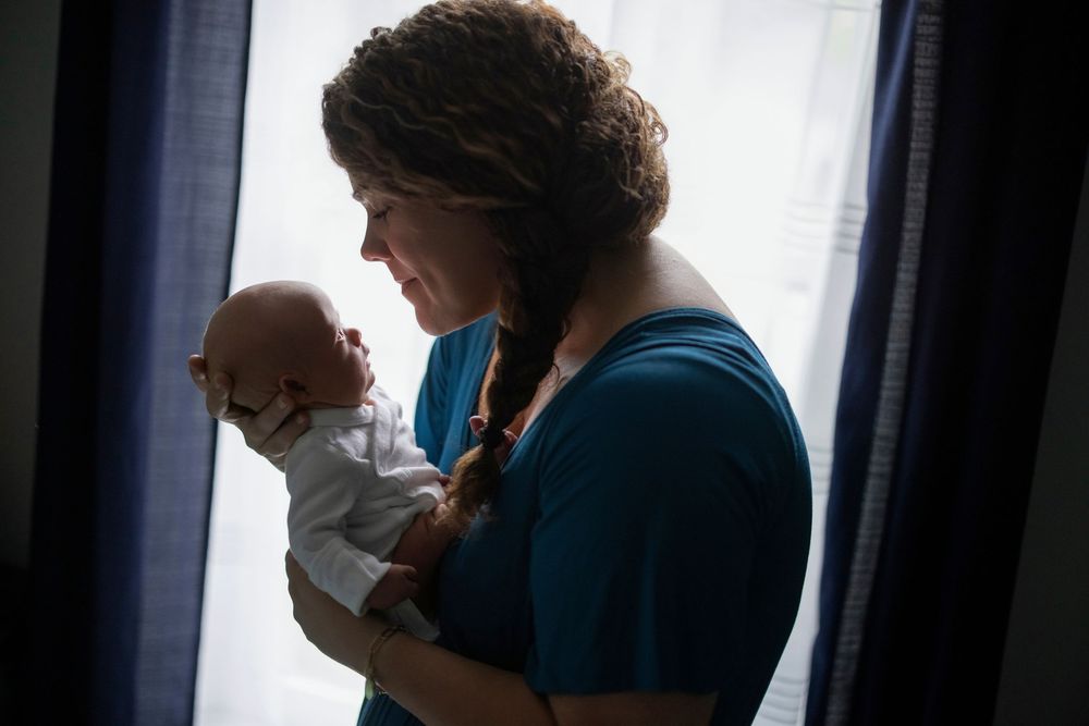 A mother tenderly holds her newborn baby near a bright window in a heartfelt portrait.