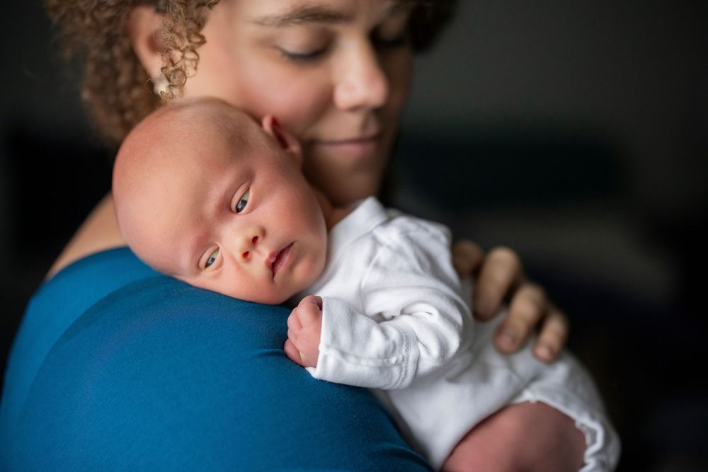 Mother tenderly holds sleeping newborn baby against her shoulder in a warm, intimate portrait.