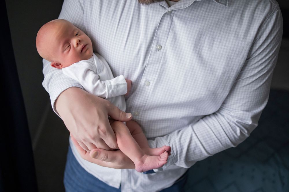 Adult cradling a sleeping newborn baby dressed in white, captured in a tender close-up portrait photo.
