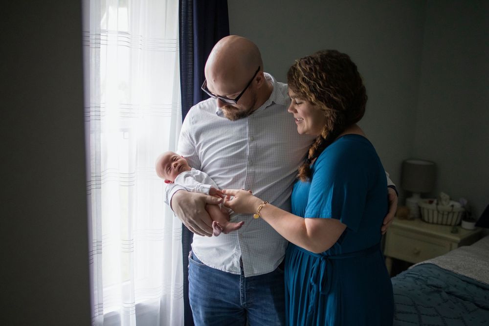 A couple tenderly holds their newborn baby by a sunlit window in a softly lit bedroom.