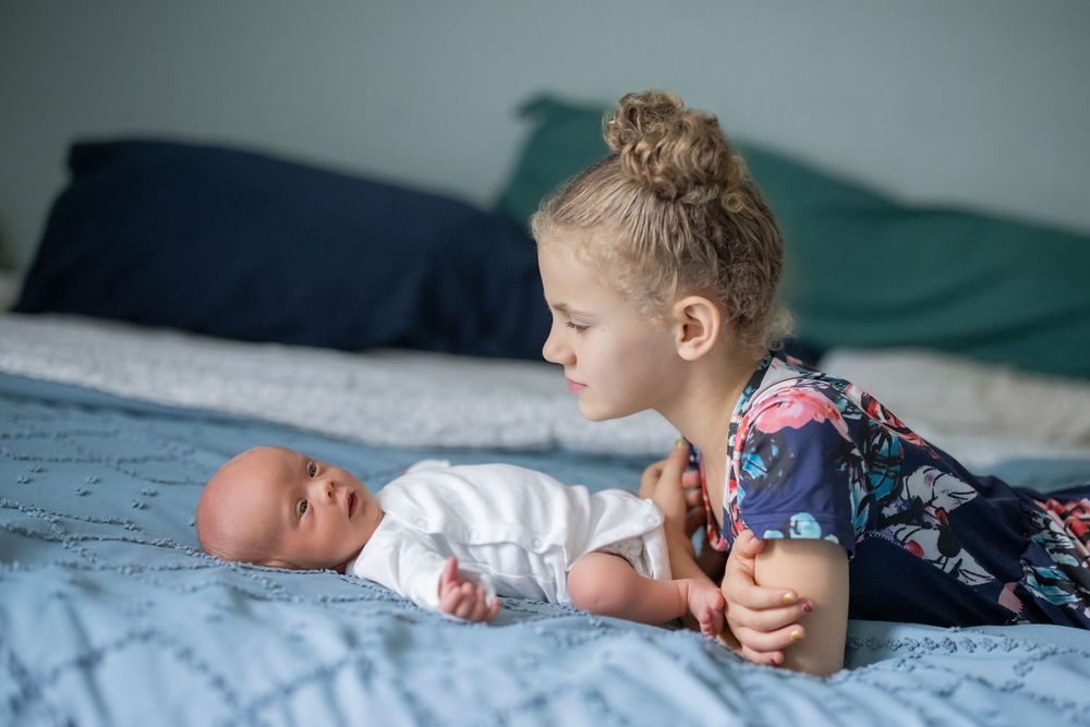 Young girl with hair bun lying on bed smiling at newborn baby sibling in white onesie.