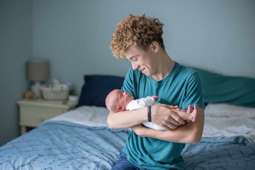 Young curly-haired man in teal shirt holding newborn baby while sitting on bed with blue bedding.