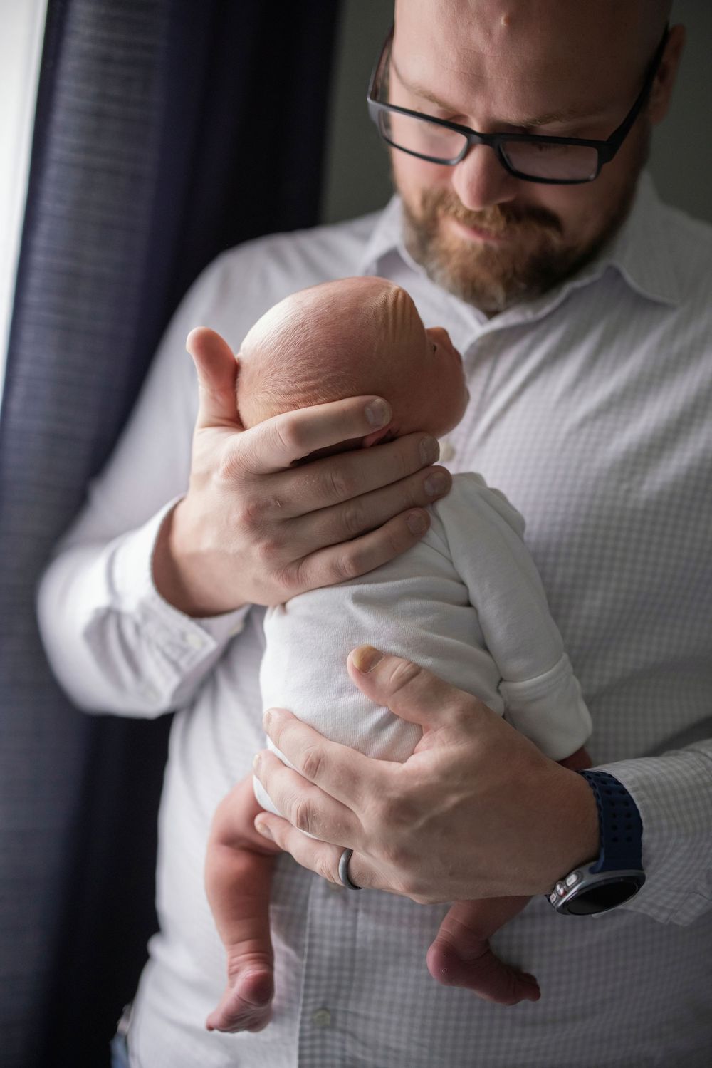 Bearded father with glasses tenderly holds newborn baby in white onesie near window.