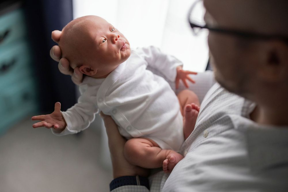 Father holding newborn baby in white onesie, infant with spread fingers in hospital setting.