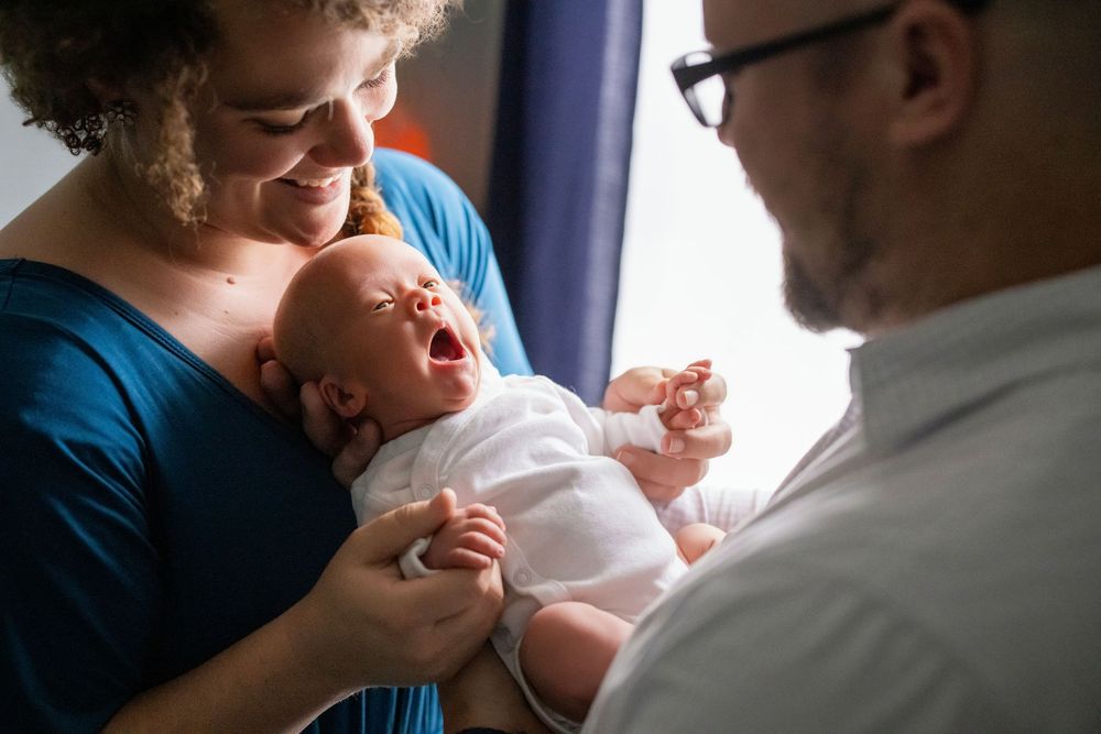 Parents smiling at their yawning newborn baby wrapped in white cloth indoors.
