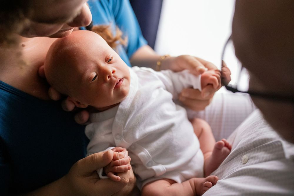 A newborn baby sleeping peacefully in a mother's arms while a doctor examines with a stethoscope.