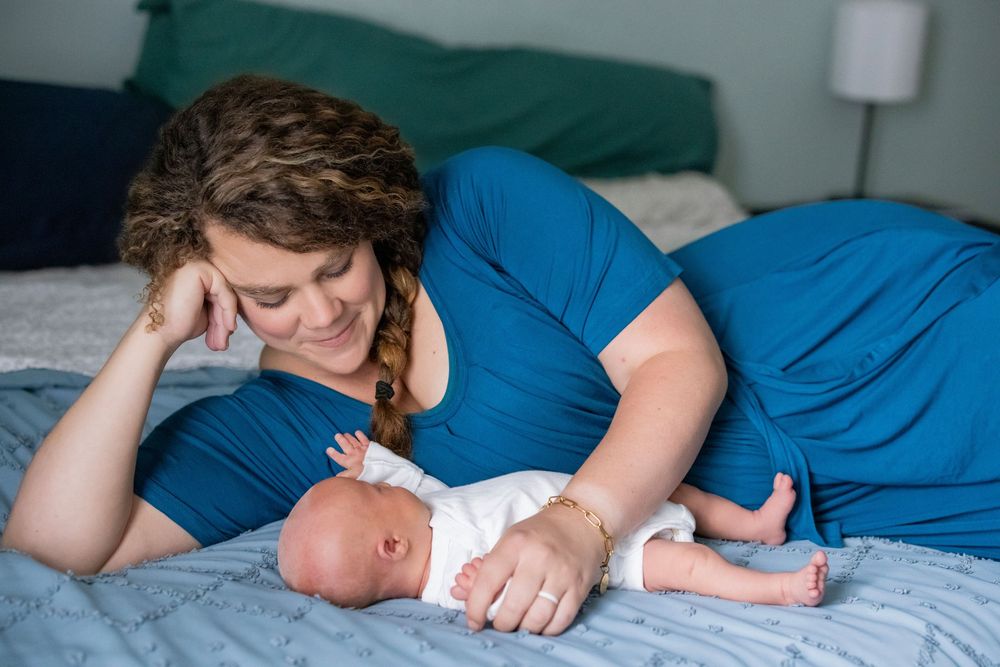 Mother in blue dress lying on bed smiling at her newborn baby.